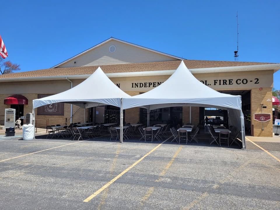 Two white high-peak tents with sidewalls set up in front of a fire company building