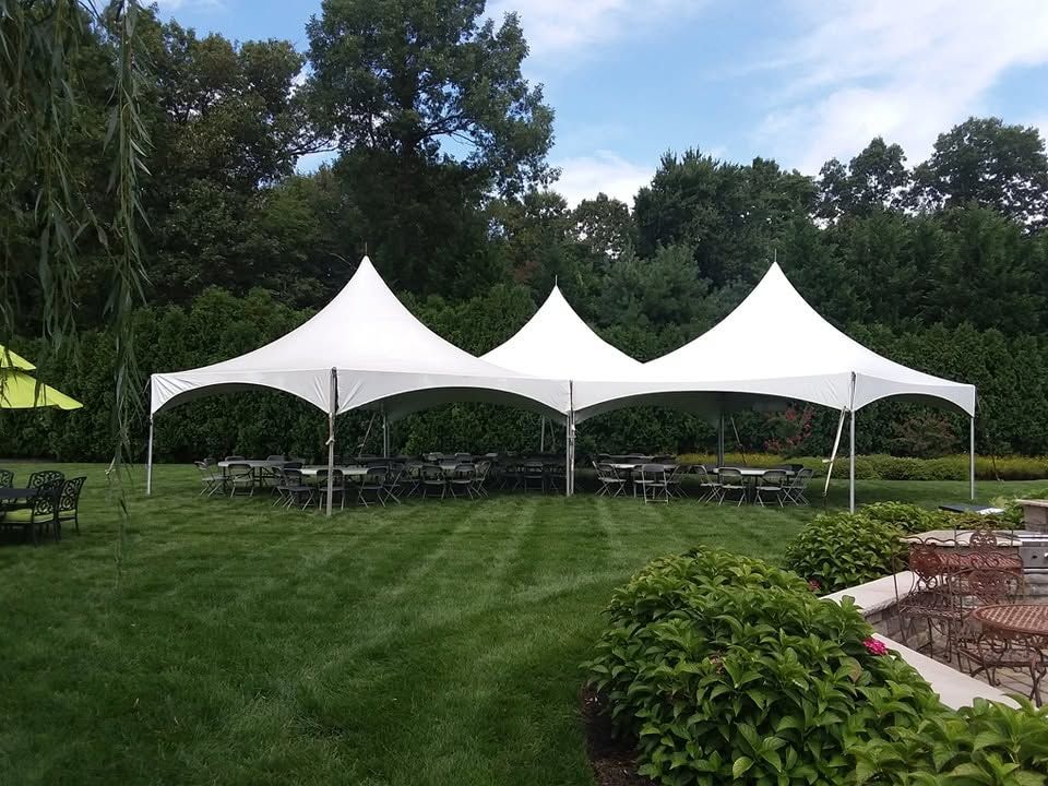 Three connected white high-peak frame tents on manicured backyard lawn with tables and chairs
